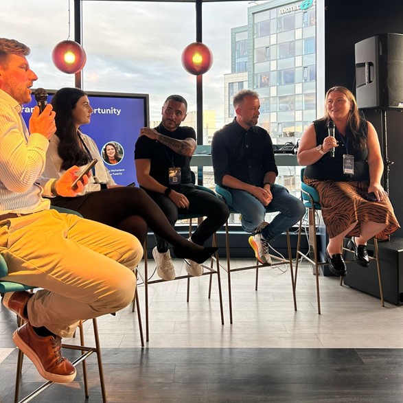 Five people sit on tall stools in a semi-circle in front of a screen - you can make out the end of the words 'your brand, your opportunity' on it. The people in the picture are, from left to right: Ewan Anderson (host), Rachael Higgins, Brian Fraser, Andrew Dobbie, and Kerry McFarlane (the four speakers). Kerry is holding a microphone and is speaking, while looking at Ewan.They are sitting in front of a window and outside you can see the MotelOne building.