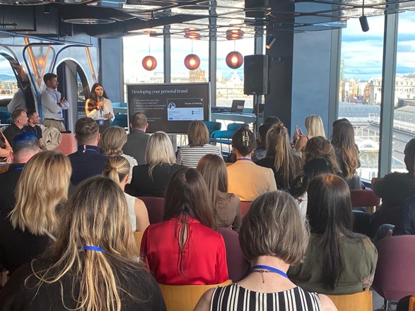 Rachael Higgins stands in front of a crowded room of attendees, discussing personal branding. She is holding a microphone as she talks. To her left is a screen, which reads 'developing your personal brand'. Out a window to the far right you can see the Glasgow skyline.