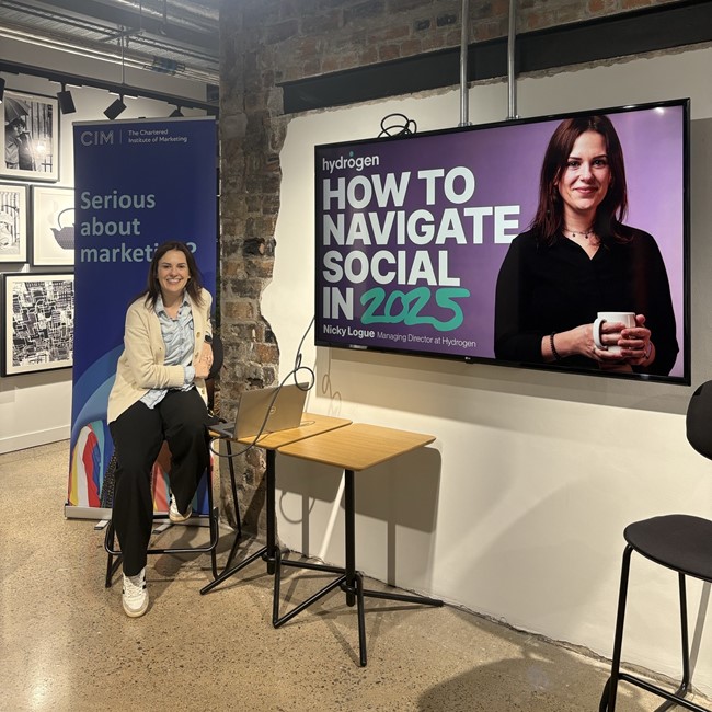 Nicky Logue smiles at the camera. Behind her is a large TV screen showing her face and the words "how to navigate social in 2025"