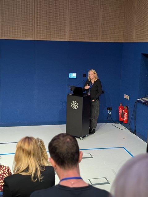 Alice Brady, a blonde woman, stands behind a lectern and gestures as she speaks to a crowd