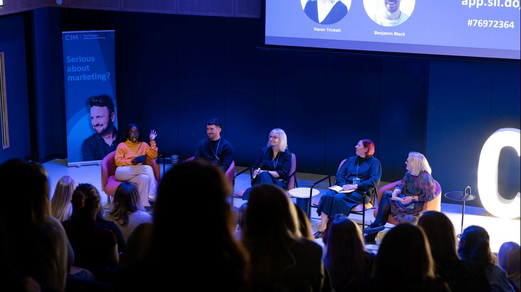 A panel of five people site on chairs in front of a crowd. Behind them is a screen and a CIM banner.