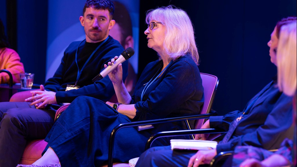 Karen Trickett, a blonde woman in glasses, holds a mic as she makes a point